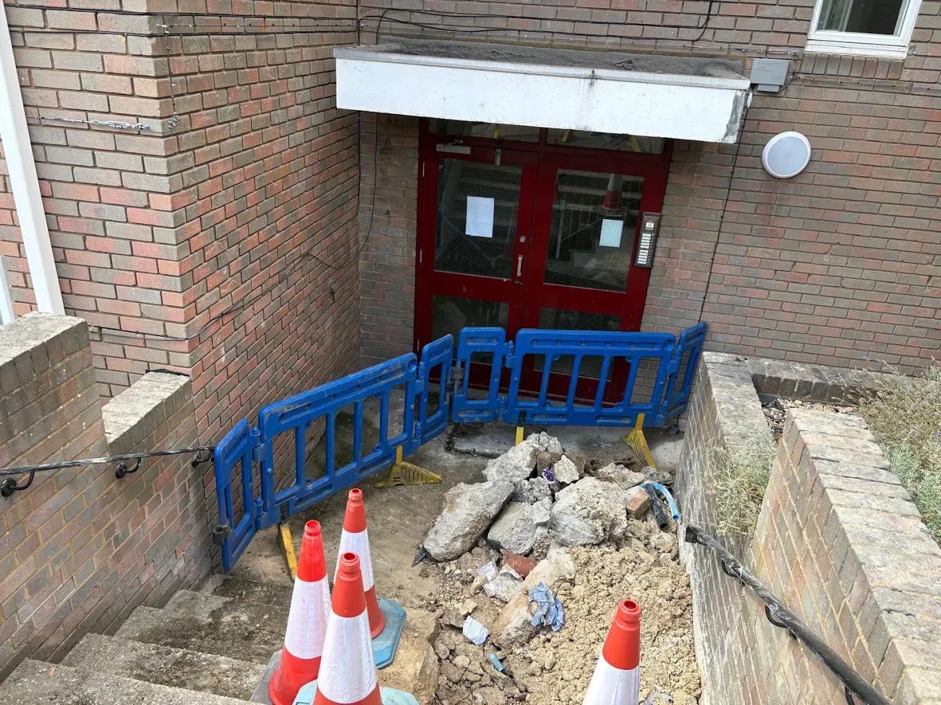 Concrete broken out at the foot of basement steps outside a block of flats, fenced off with barriers and cones
