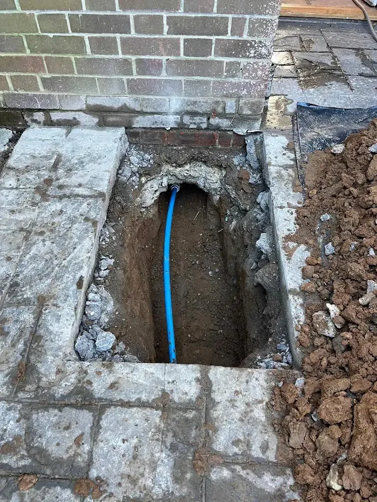 A pit dug through paving at the front step of a house, with a new blue supply pipe running into the wall