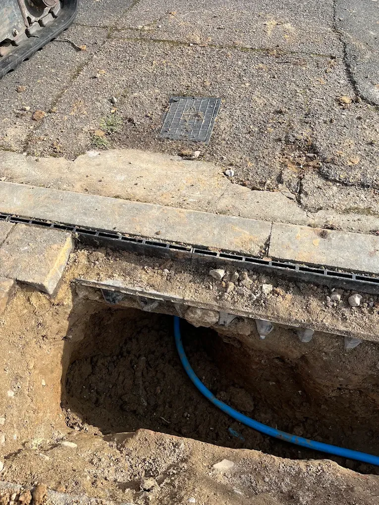 A pit at the edge of a concrete drive with new blue pipe passing beneath the drainage channel