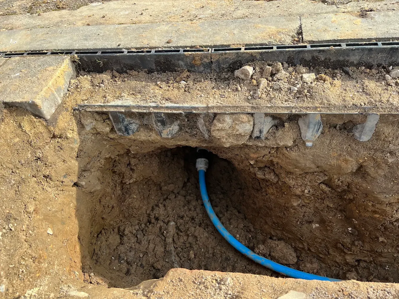 A pit dug beside a drainage channel at the mouth of a drive, with new blue pipe curving underneath it