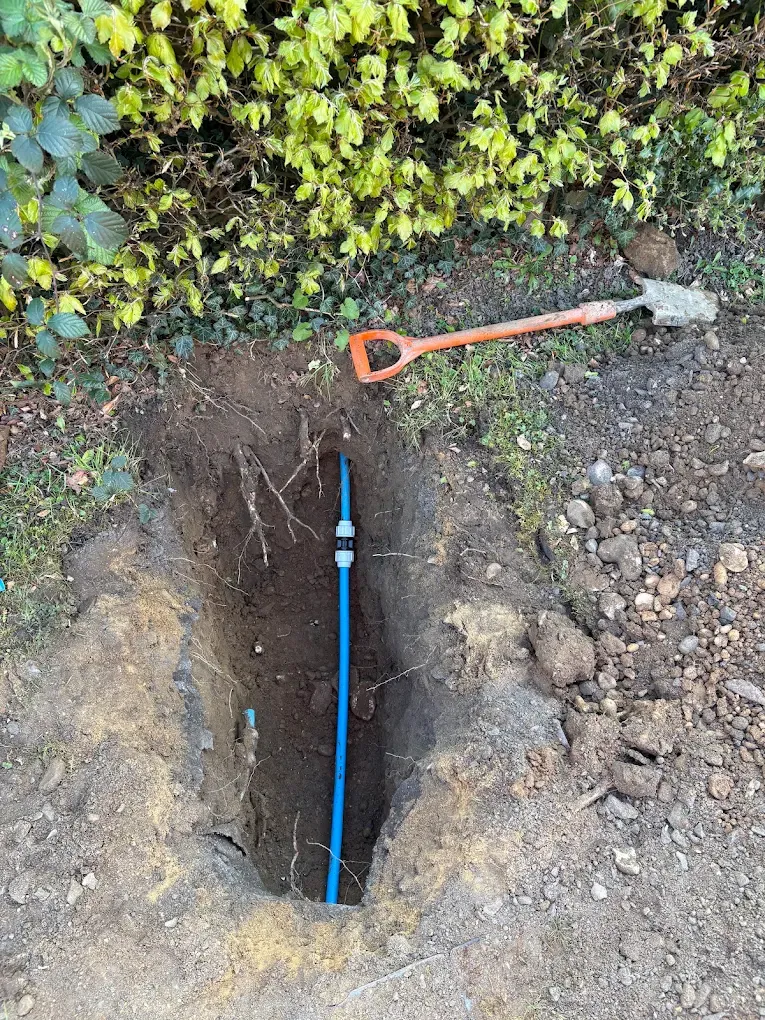 A deep narrow pit under a hedge with a length of new blue pipe running down into it and a spade resting on the edge