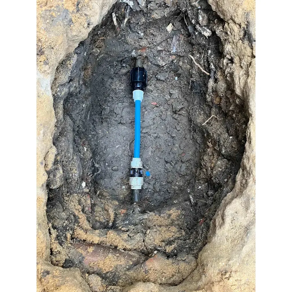 A hand-dug hole in dark clay with a short length of new blue pipe and couplings jointed into the old supply