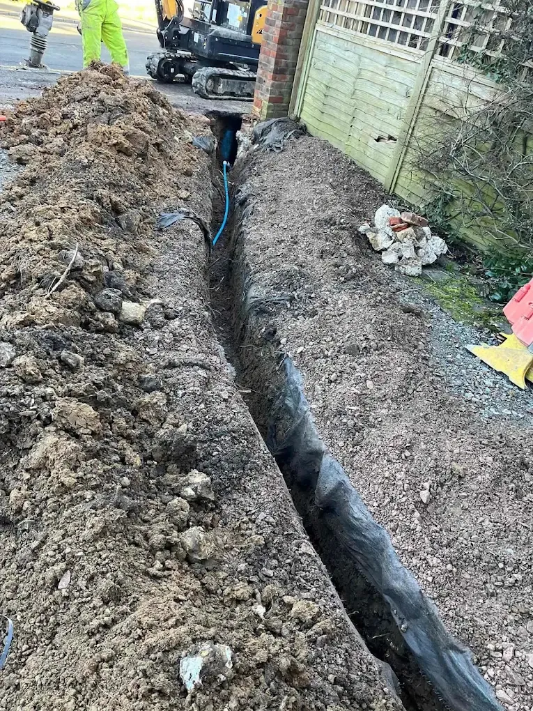 A long narrow trench beside a garden fence with new blue pipe laid in the bottom and a micro digger at the far end