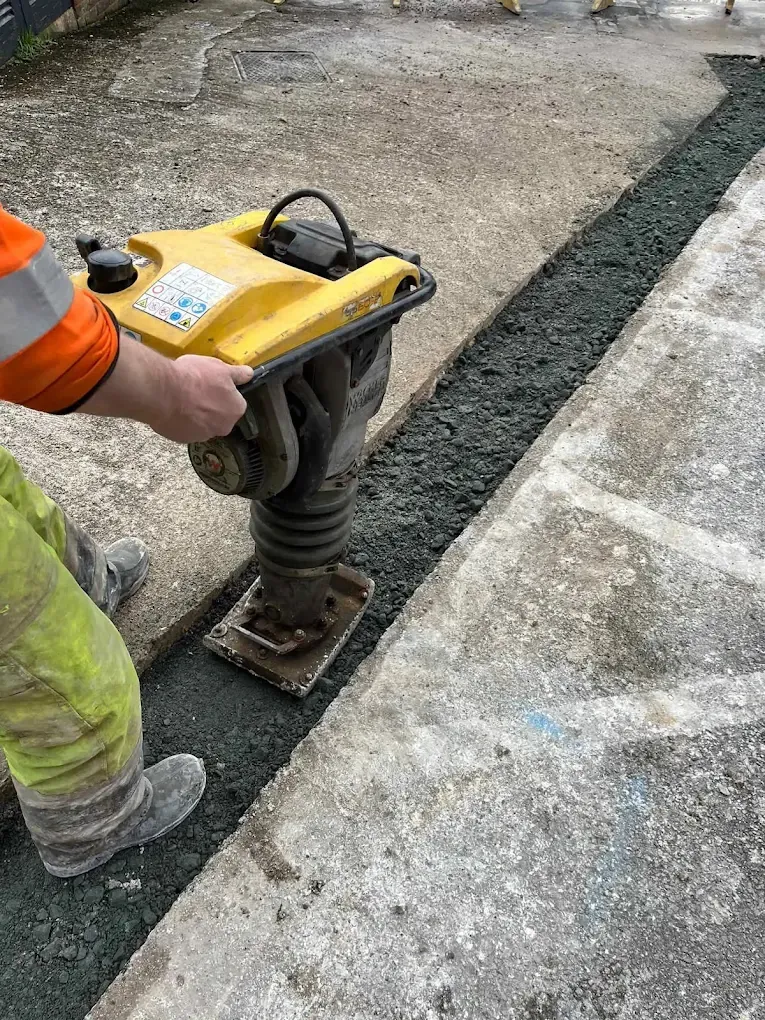A trench along a concrete path being compacted with a trench rammer before reinstatement